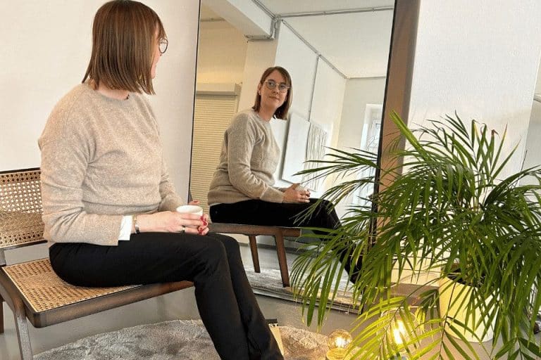 Frau sitzt auf einer Holzbank und blickt nachdenklich in einen Spiegel, umgeben von Pflanzen in einem hellen Raum.